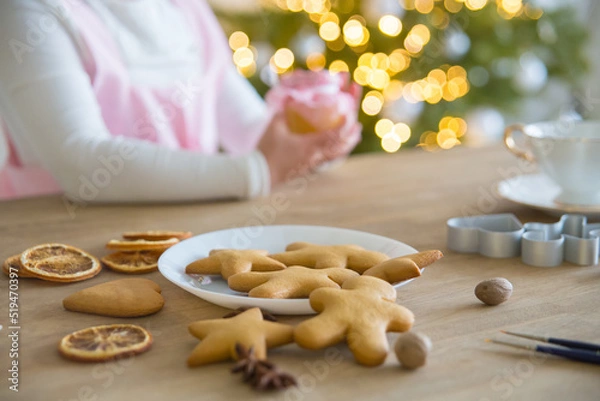 Fototapeta Christmas traditional cookies stars, hearts, men on a white plate. On the wooden table are dry oranges, anise, cookie molds and tassels. Children's hands, Christmas tree with lights in the background