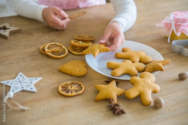 Fototapeta A child's hand takes from a white plate Christmas homemade cookies in the shape of a star, On a wooden table lie cookies gingerbread man, dried orange, anise, peenium molds and a white forfor cup