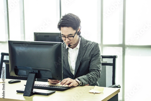 Fototapeta Young asian businessman with headphone checking updated news, texting message on mobilephone while working in office. Concept of news updated, real time working, business, financial market.