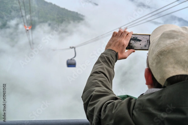Fototapeta Close-up of man taking a photo on his smartphone from cable car cabin in the mountains. Traveling concept. 