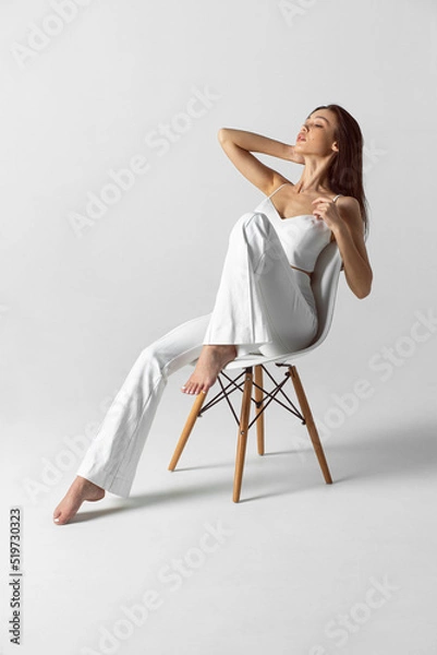 Fototapeta The full length image of a young brunette woman with makeup and hairstyle, dressed in white, sits fashioable on the chair.
