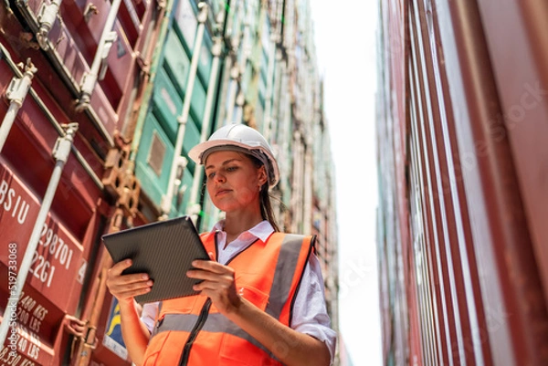 Obraz Dock manager or engineer worker in casual with vest standing in shipping container yard looking at tablet. Import and export product. Manufacturing transportation and global business concept