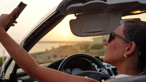 Fototapeta Attractive woman makes selfie and takes photos of the sunset from her convertible car parked in front of the beach, enjoying the sunset alone and relaxing with the view