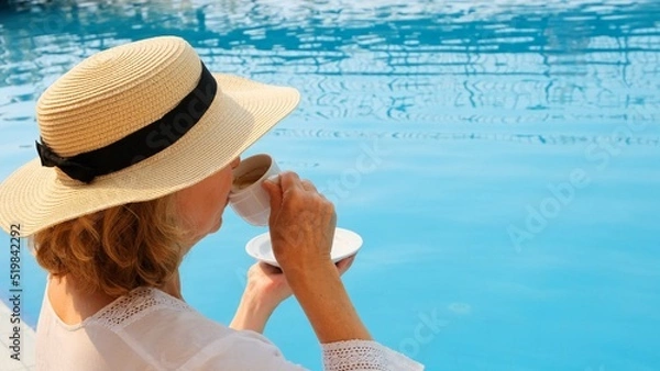 Fototapeta Close up of a woman aged 50-55 in a straw hat drinking from a cup of coffee next to a blue swimming pool, luxurious good morning, start of the day