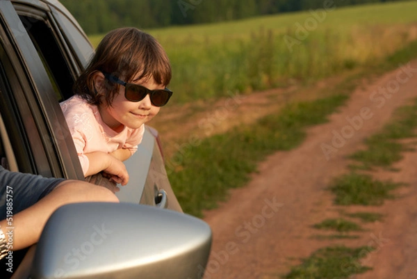 Fototapeta A fragment of a car on a field road with a smiling girl in sunglasses in the back window and motorists at the wheel. Copy space.