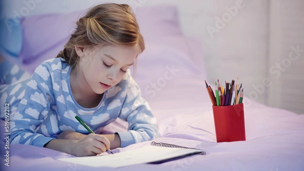 Fototapeta Child in pajama drawing on sketchbook on blurred bed in evening.