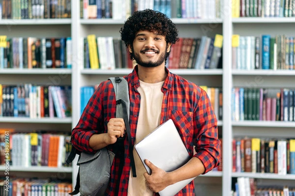 Fototapeta Happy confident indian or arabian male student of university, in stylish casual wear, with backpack and laptop, stands in a library against the background of bookshelves, looks to the camera, smiles