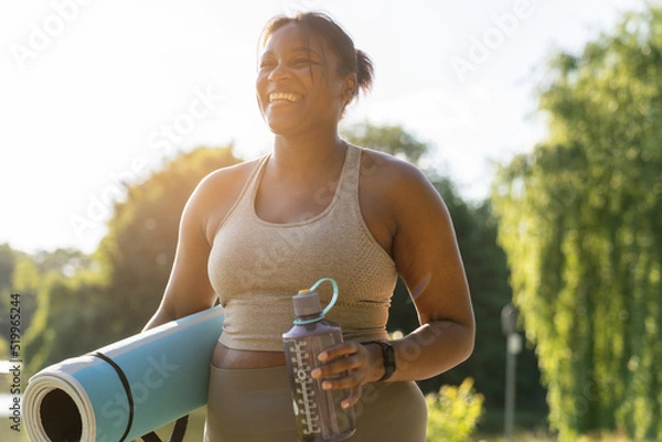 Obraz Young African American woman walking with exercise mat and bottle of water through the park in a summer day