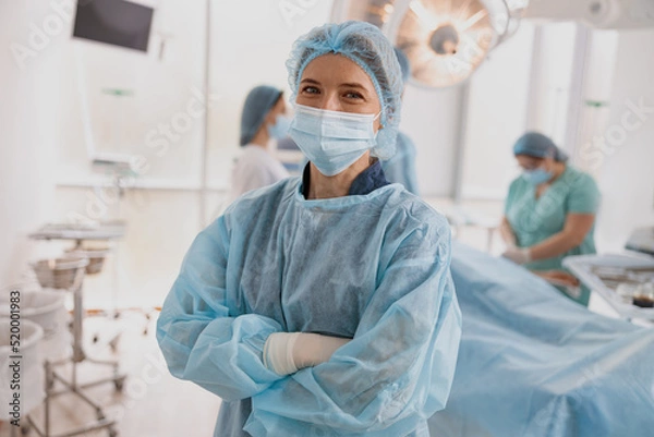 Fototapeta Female surgeon in mask standing in operating room with crossing hands, ready to work on patient