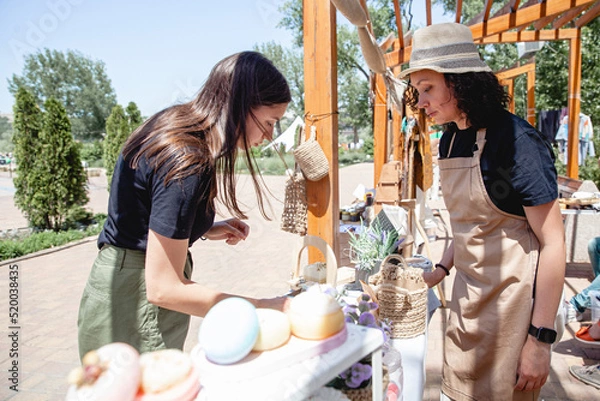 Fototapeta Young brunette girl buyer with long hair and woman seller in apron and hat on either side of the counter with goods at the street fair in summer
