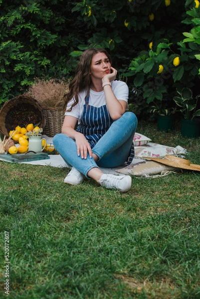 Fototapeta A girl in a striped apron and a straw hat sits on the grass near the house, holds a lemon in her hands and looks at it. Summer holiday, healthy food and ecology concept
