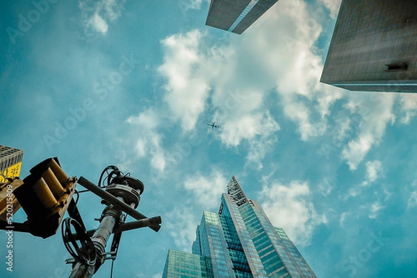 Fototapeta A low-angle view through the modern skyscrapers with the typical traffic yellow lights, jet plane, and cloudy turquoise blue sky in the background. 
Manhattan, New York City, USA. 