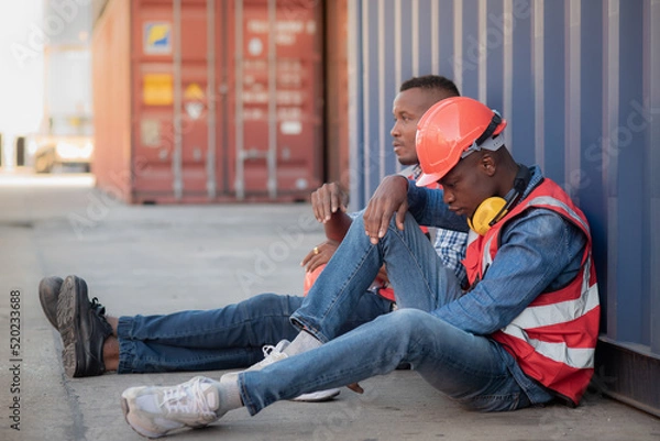 Obraz 2 African lack man worker wearing safety uniform workwear and sitting on the floor leaning against the container, there is stressed and no work to do, economic recession due to inflation.