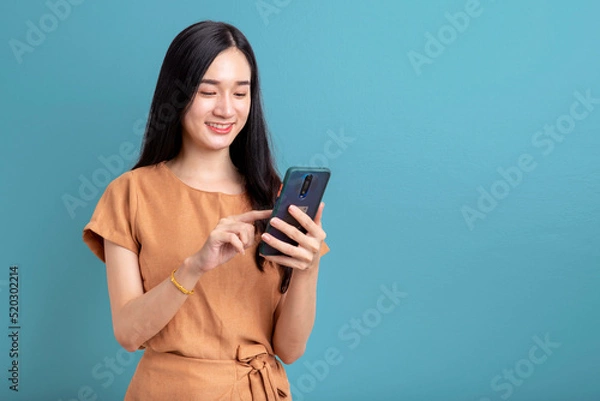 Fototapeta Happy Portrait smiling young asian woman using smartphone isolated on blue background