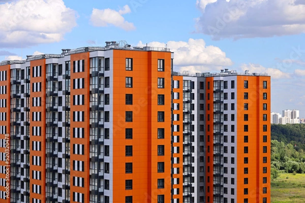 Fototapeta New residential buildings with orange lining on background of green park and blue sky with white clouds. House development, high-rise construction in ecologically clean area