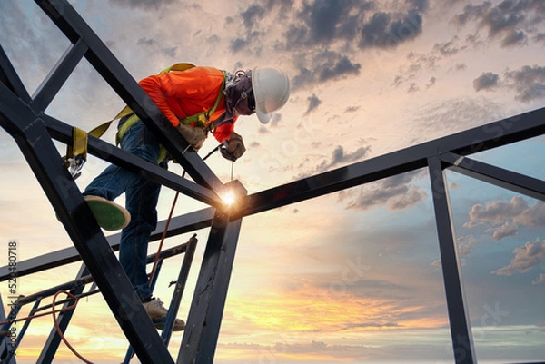 Fototapeta A welder is welding steel on a steel roof truss. Working at height equipment. Fall arrestor device for worker with hooks for safety body harness. Worker in construction site.