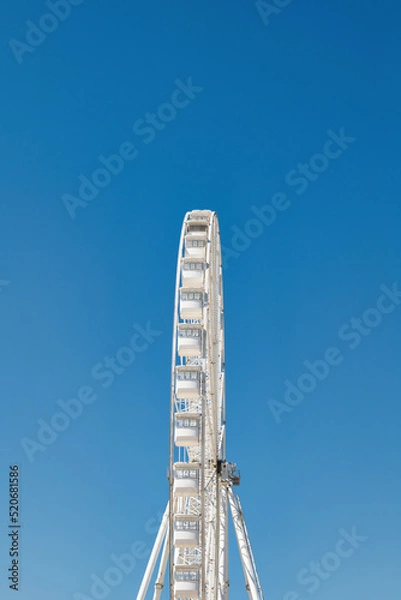 Obraz Side view of a tall white metal Ferris Wheel in front of a clear blue sky. Summer vacation advertisement or presentation background. 