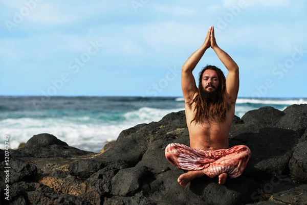 Fototapeta Rasta boy with beard doing yoga posture sitting with his hands together on his head. by the sea.
