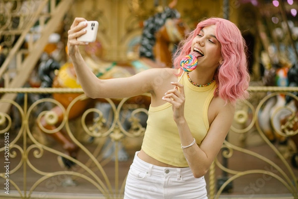Fototapeta Charming girl playing with two colorful lollipops. Girl with pink hair posing with candies. carousel in the background. Rest in the amusement park