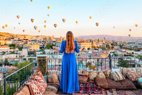 Fototapeta young woman in blue dress stands on terrace of hotel in Goreme Cappadocia and looks at hot air balloons rising into sky, concept  of must see travel destination, bucket list trip