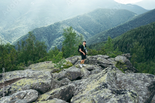 Fototapeta A guy tourist with a backpack stands on a stone on top of a mountain against the background of beautiful views.