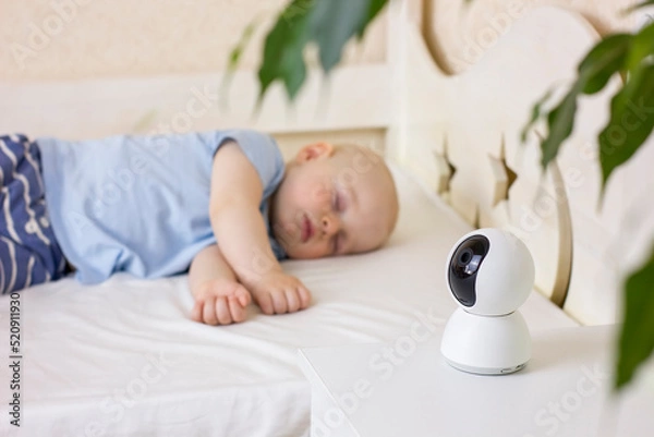 Fototapeta Cute little baby boy sleeping on bed at home with baby monitor camera.