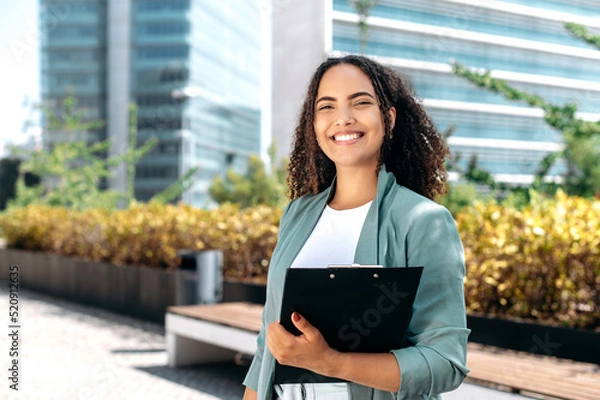 Obraz Elegant confident young mixed race woman with curly hair, formally dressed, stands outdoors against the background of the business center, holding a folder with documents, looks at camera, smiles