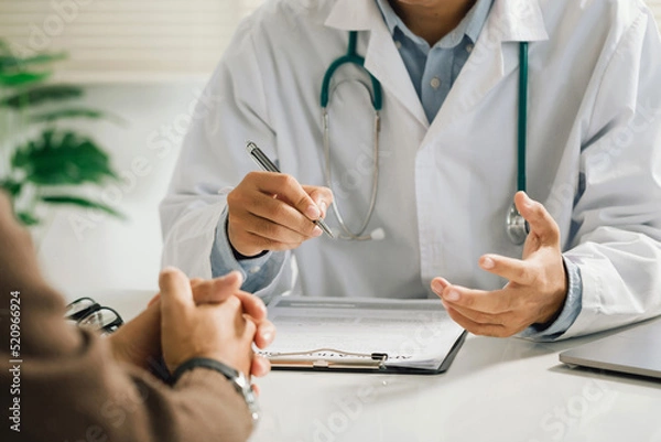 Obraz A professional physician in a white medical uniform talks to discuss results or symptoms and gives a recommendation to a male patient and signs a medical paper at an appointment visit in the clinic.