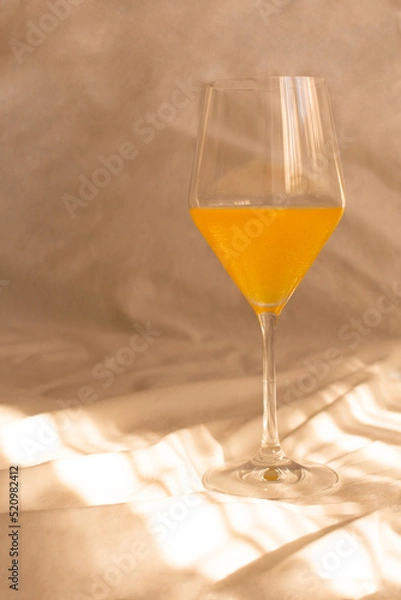 Fototapeta Orange juice in an elegant wine glass on a sunlit white sheet. Romantic aesthetic. Natural sunlight and shadows.