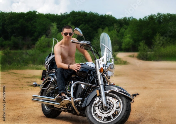 Fototapeta A young man of athletic build is sitting on a motorcycle and posing in front of the camera.