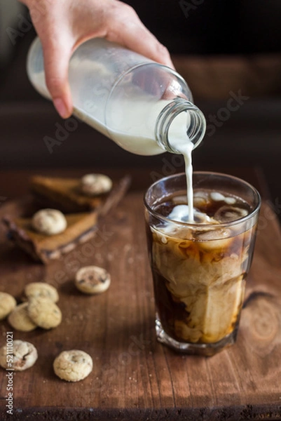 Fototapeta A woman's hand holds a bottle of milk and pours it into a glass of cold coffee. on the table in the background lies chocolate and biscuit cookies