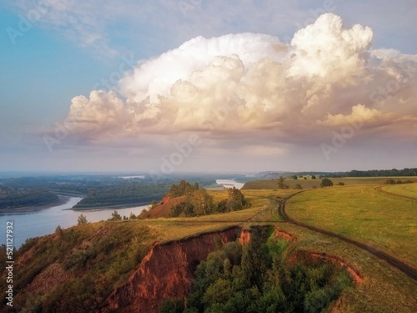 Obraz In large clouds, the light of the setting sun and the warm evening light flood the trees on the hill, the clay mountain and the reflection of the clouds in the water. river Ob.