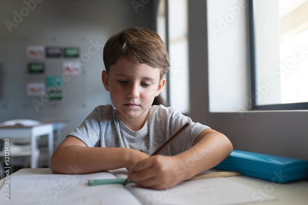 Fototapeta Caucasian elementary schoolboy writing on book while sitting at desk in classroom