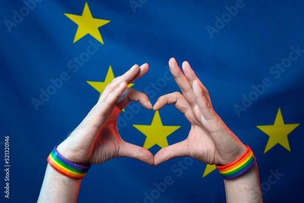 Fototapeta Men's hands with bracelets with LGBT symbols folded a heart from their fingers against the background of the European Union flag