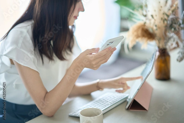 Fototapeta PC画面を前に、スマートフォンを操作する女性