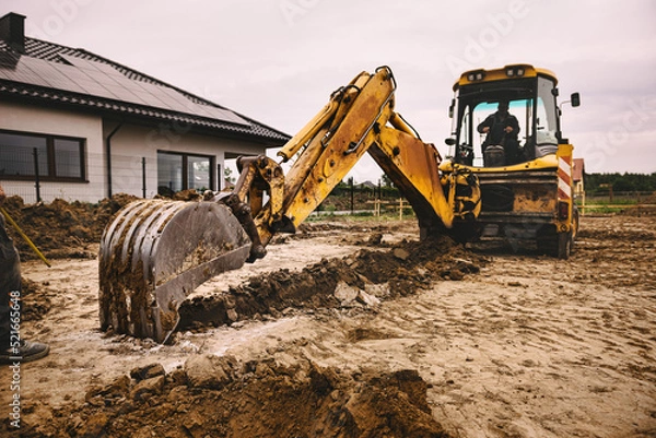 Obraz Excavator working at house construction site - digging foundations for modern house. Beginning of house building. Earth moving and foundation preparation.
