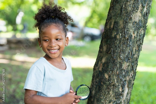 Fototapeta Smiling African American little girl has fun holding magnifying glass to explore and look bugs on the tree between learning beyond the classroom. Education outdoor concept.