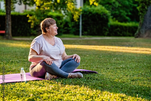 Fototapeta Relaxed senior plus size woman with earphones sitting on yoga mat on in park outdoors resting after exercises at warm sunny summer morning