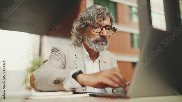 Fototapeta Mature businessman with beard in eyeglasses, user on laptop computer and taking notes, sitting in outdoor cafe. Successful man sitting at table in cafe in nature Mobile office freelancer