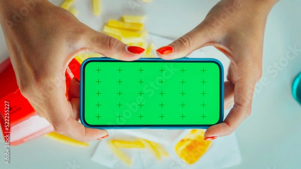 Fototapeta Female hands hold horizontal smartphone with chroma key green screen over table at cafe. French fries and burger at cafe, woman use mobile phone with mockup. Use smartphone app top view