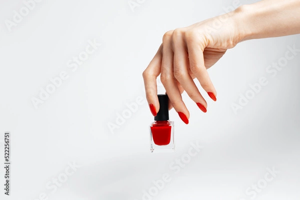 Fototapeta Close-up of female hand with red manicure holding a glitter nail polish of red color on white background.