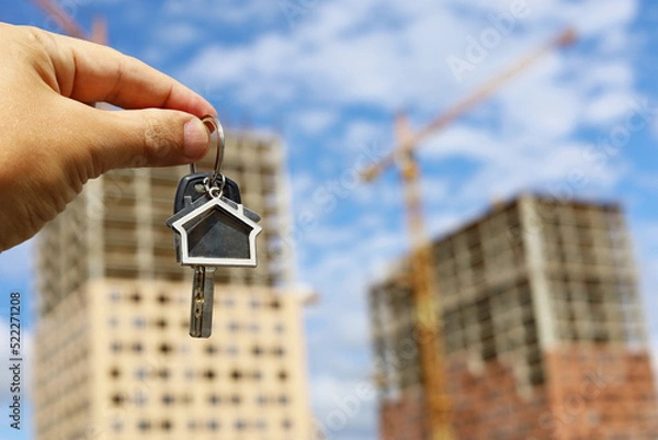 Fototapeta Keychain in house shape and key in male hand on background of construction cranes and new buildings. Real estate agent, concept of buying apartment with a mortgage
