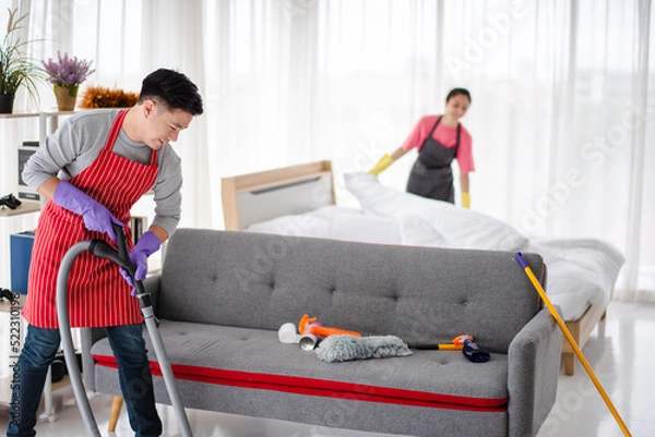 Fototapeta Young Asian family, Husband and wife are cleaning the house together. he is vacuuming,  using a vacuum cleaner, and the wife is wearing the same apron but is black. She is changing new blankets