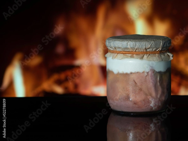 Fototapeta A jar of home-made canned meat with a parchment cover on a black surface in the background of a fireplace fire