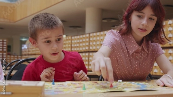 Fototapeta 4K. Children play a board game in the library, sitting at the table.