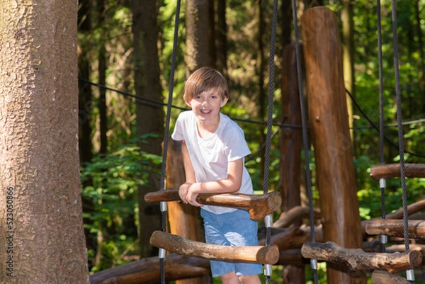 Fototapeta happy boy in the camp climbs the rope complex, overcomes obstacles in the summer in the park