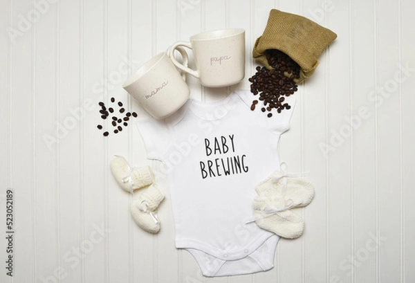 Fototapeta Baby Announcement. Baby Brewing written on a onesie with booties Coffee mugs and Spilled Coffee Beans. Flat lay on white beadboard with copy space.
