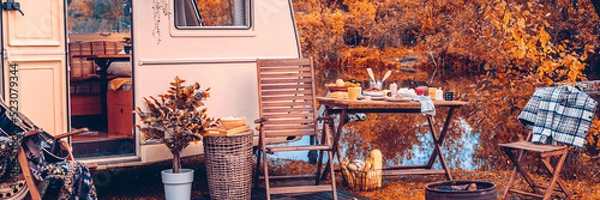 Fototapeta trailer of mobile home or recreational vehicle stands on shore of pond in camping in autumn near table set, concept of family local travel in native country on caravan or camper van and camping life