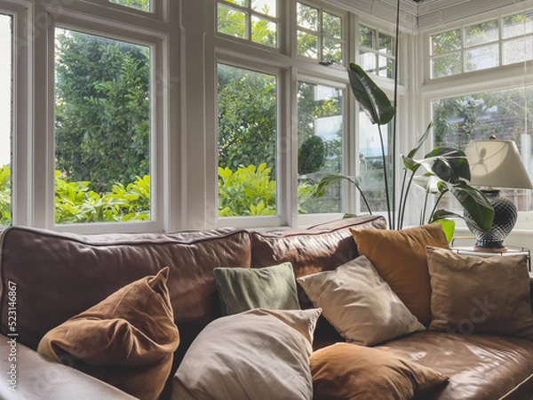 Obraz A brown leather sofa with scattered pillows, typical windows and a strelitzia plant in atypical British home. Interior decor.