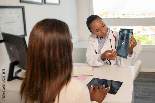 Fototapeta Confident doctor showing x-ray picture of lungs and explaining diagnosis to patient. Young African American pulmonologist consulting woman in clinic. Medical consultation, pulmonology concept